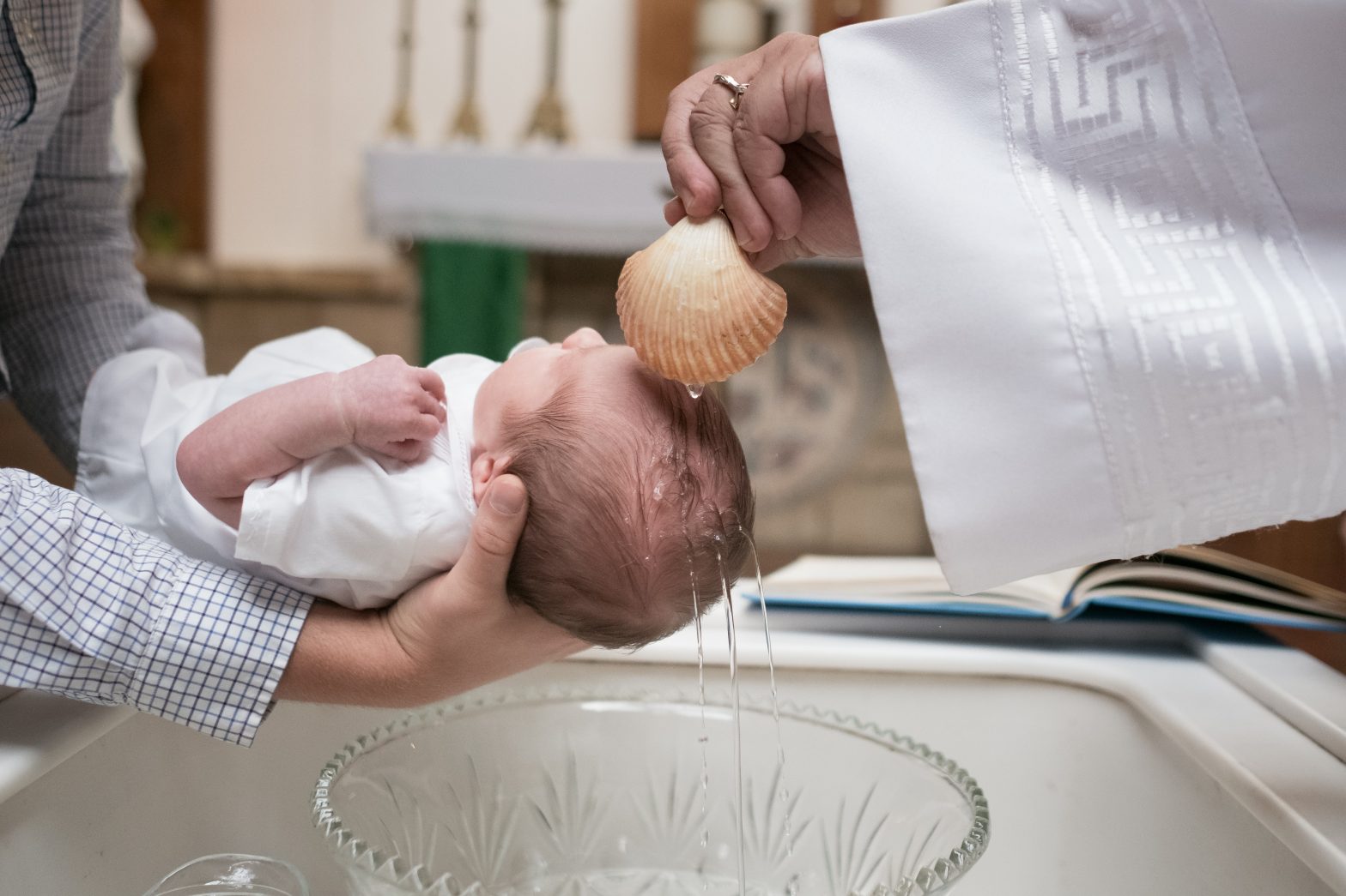 A baby being held over a font and water is poured onto their head from a shell
