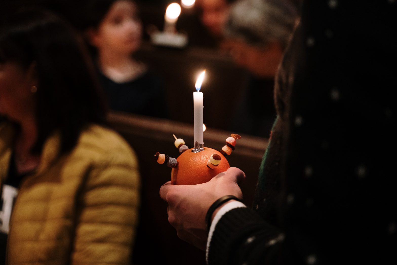 A hand holding a Christingle orange with a lit candle on top and sweets skewered around the candle