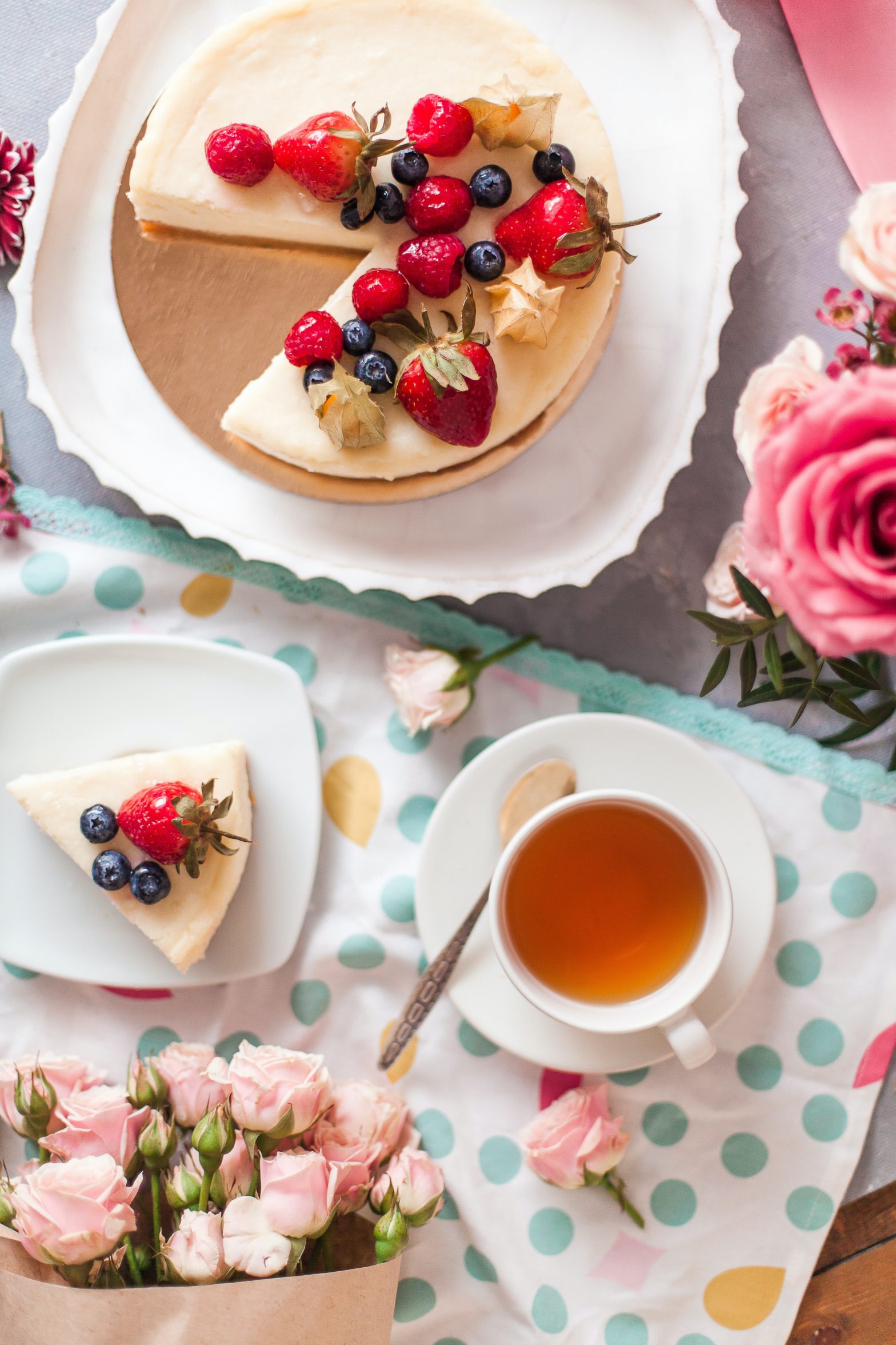 A cup and saucer of tea with a cheesecake topped in fruit, surrounded by flowers.